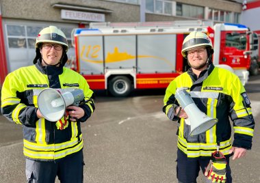 Zwei Feuerwehrleute mit Megafonen stehen vor der Feuerwehrwache.