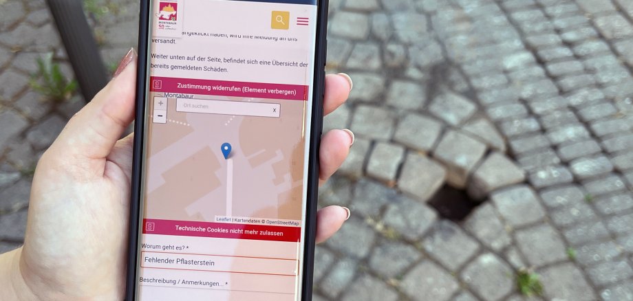 Eine Hand hält ein Smartphone über einem Loch im Gehweg. Auf dem Display ist der rlpDirekt-Schadensmelder zu sehen.