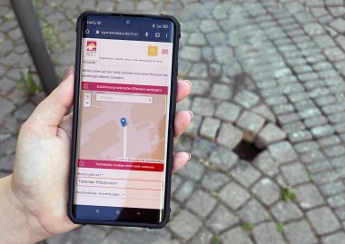 Eine Hand hält ein Smartphone über einem Loch im Gehweg. Auf dem Display ist der rlpDirekt-Schadensmelder zu sehen.