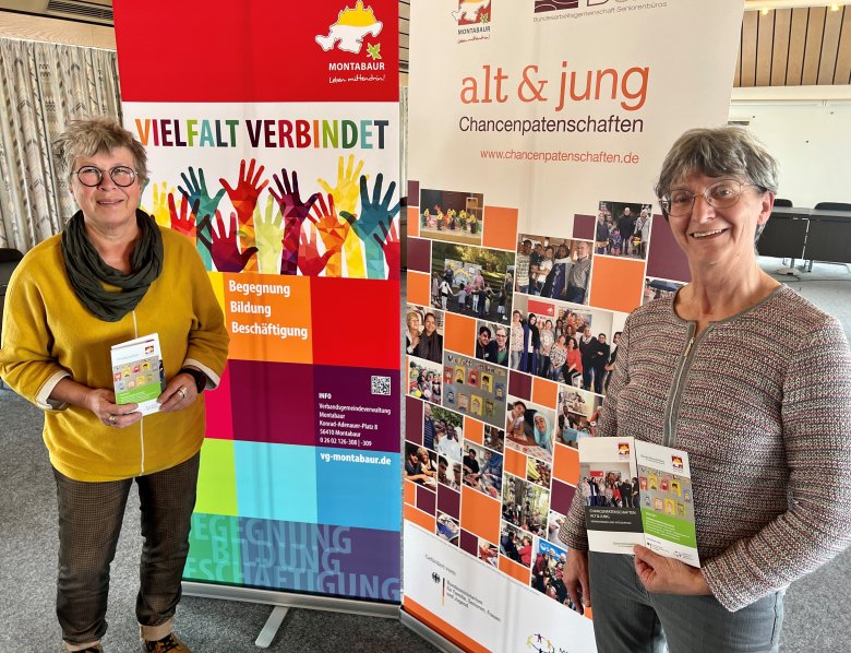 Judith Gläser und Marie-Theres Wagner stehen vor den Werbebannern des Generationenbüros und präsentieren die Flyer zum Projekt „Chancenpatenschaften Alt & Jung“.  