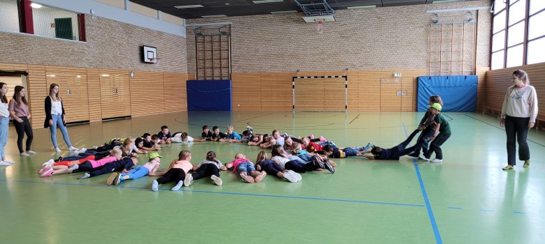 Kinder liegen auf dem Boden und bildenen einen Kreis. Zwei Kinder ziehen ein Kind an den Füßen aus dem Kreis.