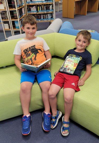 Zwei Jungen sitzen auf einer grünen Couch, einer liest in einem Buch, der andere hat es sich auf den Bauch gelegt.