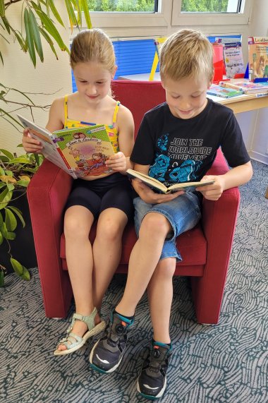 Ein Mädchen und ein Junge sitzen gemeinsam auf einem roten Sessel und lesen jeder in einem Buch