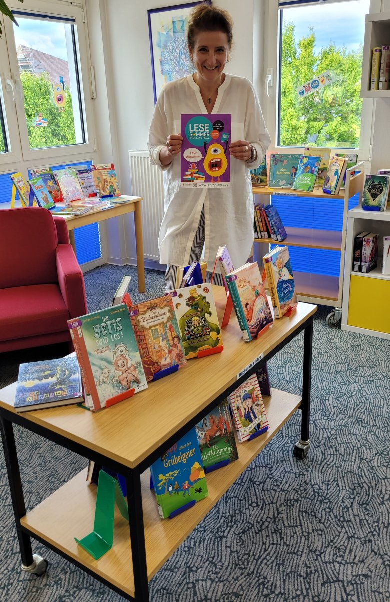 Eine Frau steht vor einem Büchertisch und hält ein Plakat mit der Aufschrift Lesesommer in der Hand