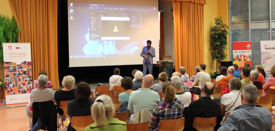 Das Bild zeigt Publikum und einen Redner vor einer Präsentationswand