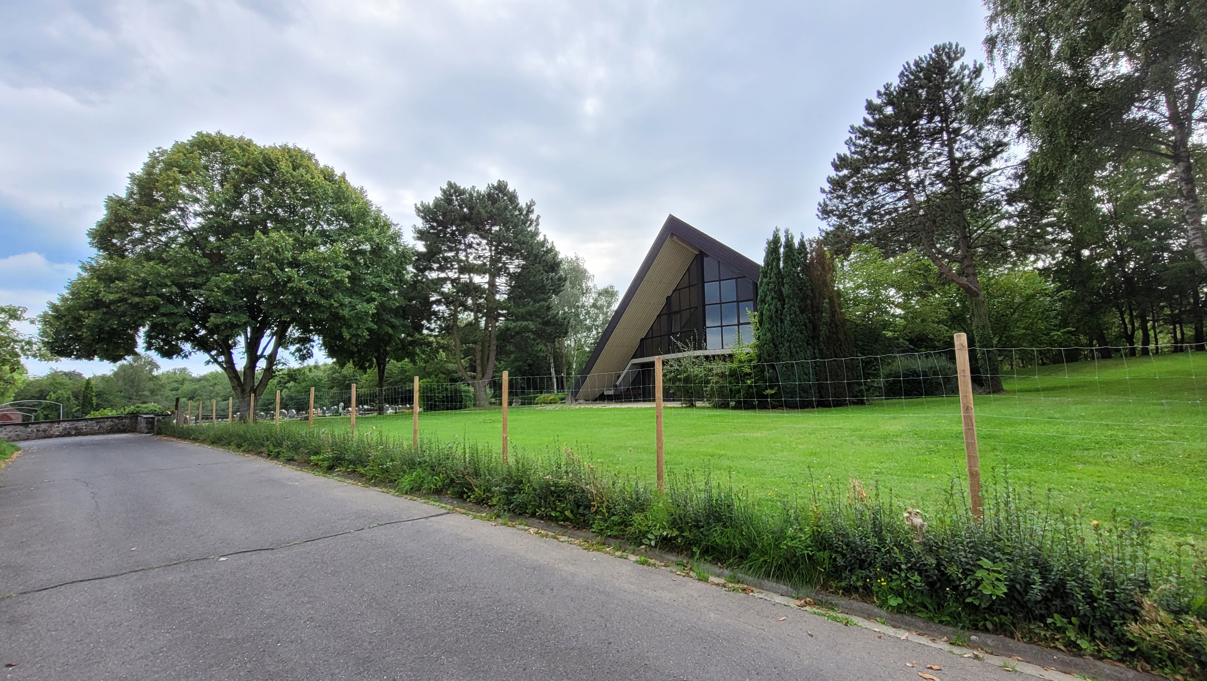 Dreieckige Gebäude auf einer grünen Wiese, umringt von Bäumen, im Vordergrund eine geteerte Straße, im Hintergrund sind die Grabsteine des Friedhofs zu erkennen