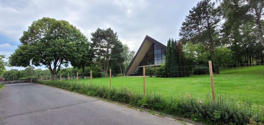 Dreieckige Gebäude auf einer grünen Wiese, umringt von Bäumen, im Vordergrund eine geteerte Straße, im Hintergrund sind die Grabsteine des Friedhofs zu erkennen