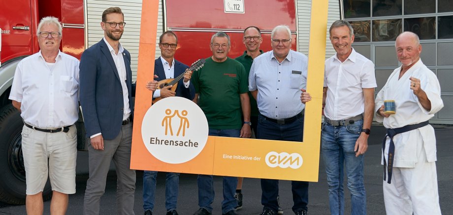 8 Männer stehen vor einem historischen Feuerwehrauto, zwei halten einen orangefarbenen Bilderrahmen mit der Aufschrift Ehrensache evm in der Hand