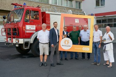 8 Männer stehen vor einem historischen Feuerwehrauto, zwei halten einen orangefarbenen Bilderrahmen mit der Aufschrift Ehrensache evm in der Hand