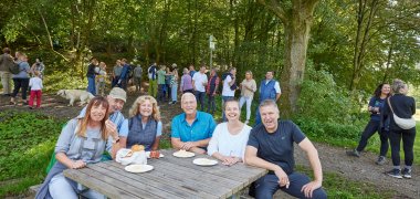 Sechs Menschen sitzen um einen Tisch, im Hintergrund sind mehr Menschen zu sehen