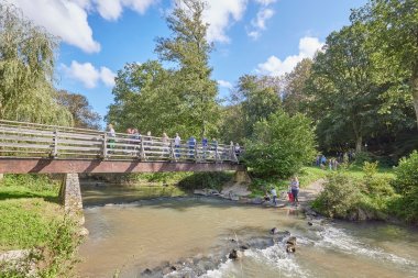 Menschen laufen über eine Brücke, die über einen Bach führt, Kinder spielen am Wasser