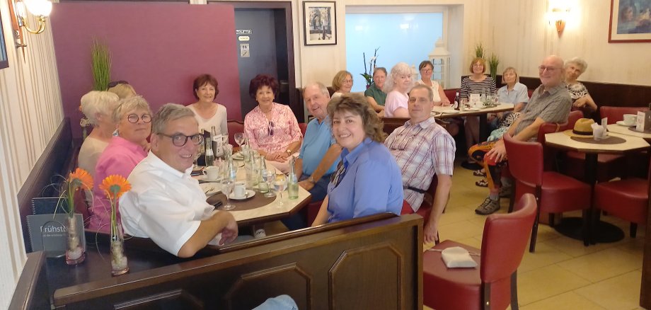  Eine Gruppe von etwa 16 Personen sitzt in einem Café an mehreren zusammengeschobenen Tischen und lächelt in die Kamera. Sie sind Teil einer Feier für den ersten Geburtstag der Initiative "PROaktiv 60+". Die Atmosphäre ist freundlich und entspannt, die Tischdekoration umfasst orangefarbene Blumen und Getränkeflaschen.