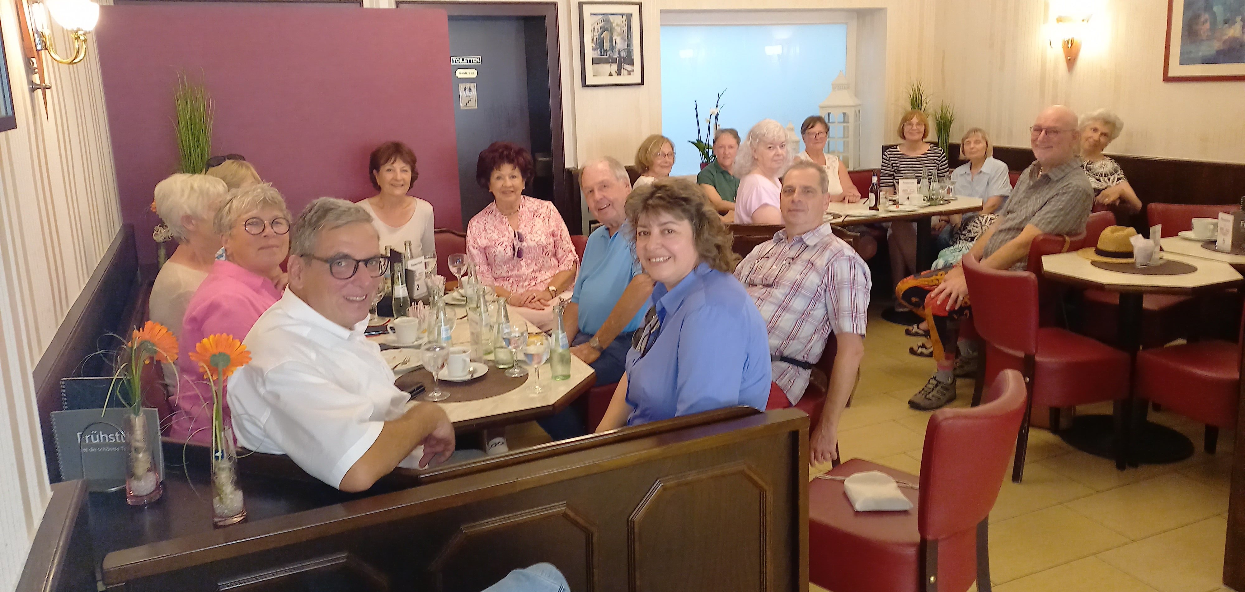  Eine Gruppe von etwa 16 Personen sitzt in einem Café an mehreren zusammengeschobenen Tischen und lächelt in die Kamera. Sie sind Teil einer Feier für den ersten Geburtstag der Initiative "PROaktiv 60+". Die Atmosphäre ist freundlich und entspannt, die Tischdekoration umfasst orangefarbene Blumen und Getränkeflaschen.