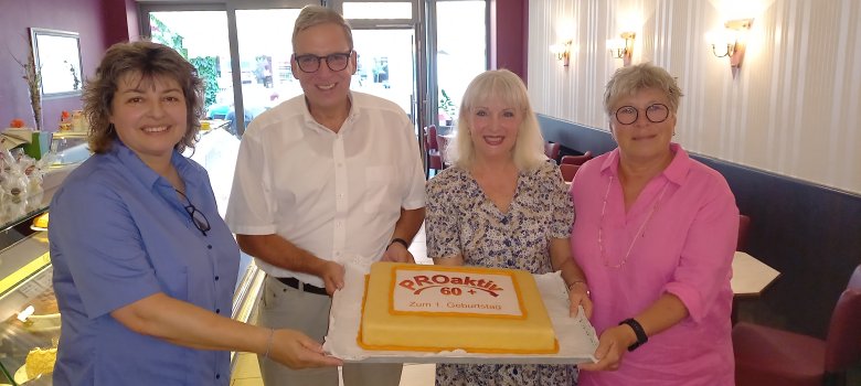 Vier Personen lächeln und halten gemeinsam einen Kuchen mit der Aufschrift "PROaktiv 60+ Zum 1. Geburtstag". Sie stehen in einem gemütlichen Café mit rot-weißen Wänden und gedeckten Tischen.