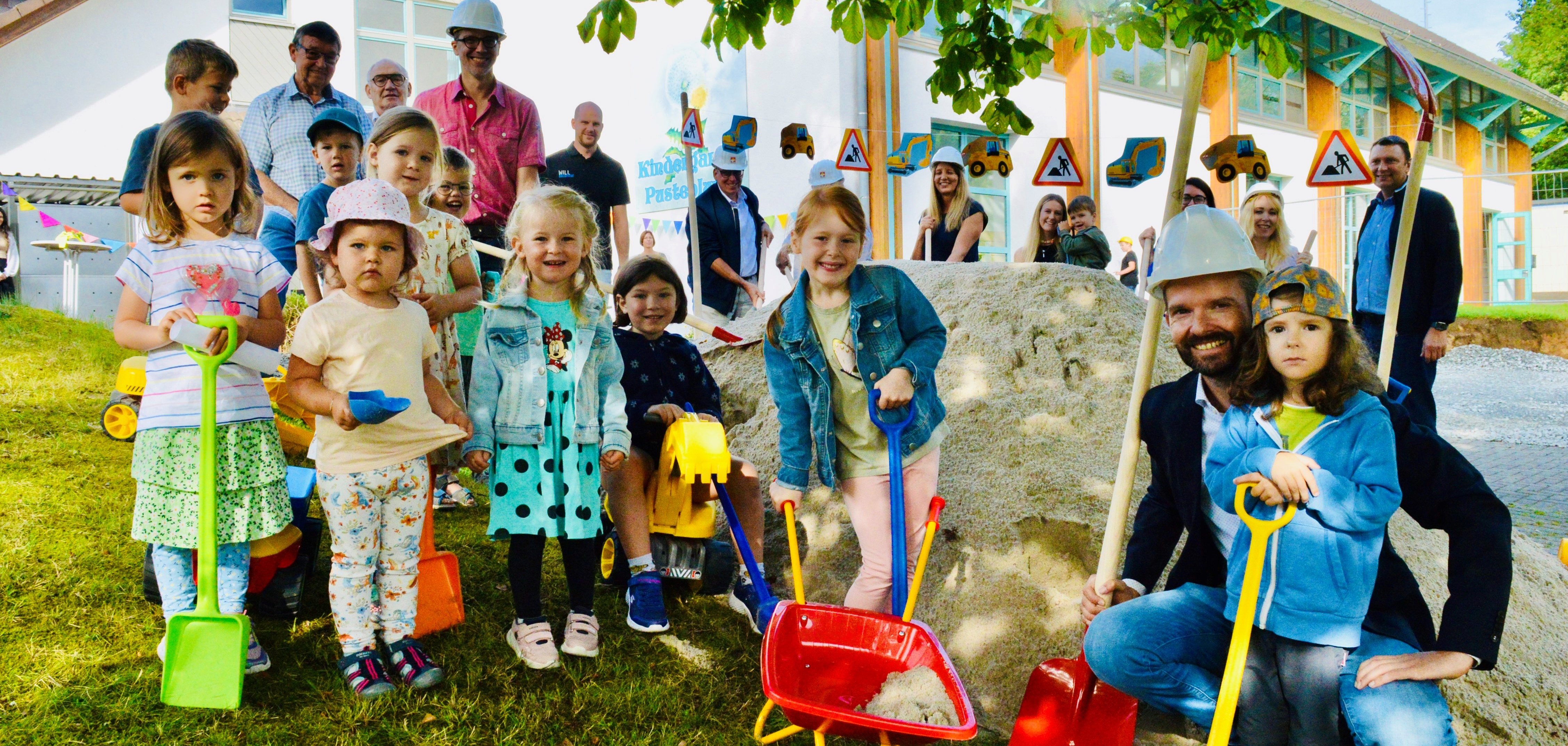 Eine Gruppe von Kindern und Erwachsenen posiert fröhlich beim Spatenstich vor der Kita Pusteblume im Freien. Die Kinder tragen bunte Kleidung und halten Schaufeln und eine Schubkarre, während einige Erwachsene im Hintergrund Bauhelme tragen. Im Hintergrund ist die Kita mit Baustellenschildern und Spielzeug-Baumaschinen zu sehen.