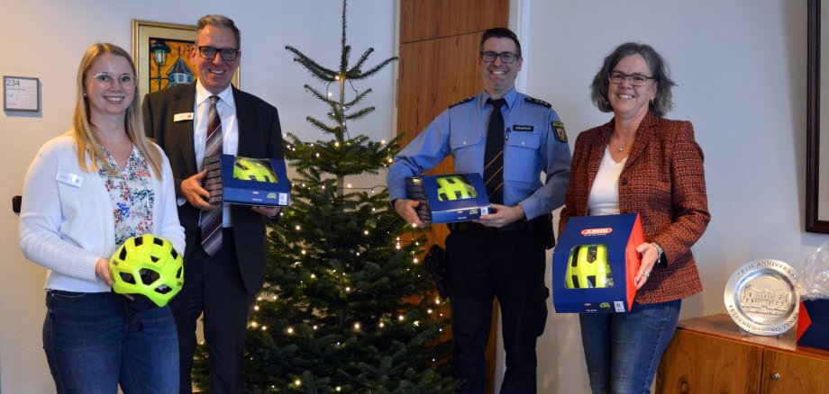 Ein Gruppenfoto mit vier Personen vor einem geschmückten Weihnachtsbaum. Alle halten leuchtend gelbe Fahrradhelme, teilweise in Verpackungen, während weitere Helme am Boden gestapelt sind. Die Szene scheint eine Spenden- oder Geschenkübergabe zu sein, möglicherweise für eine Verkehrsschule oder eine ähnliche Initiative.