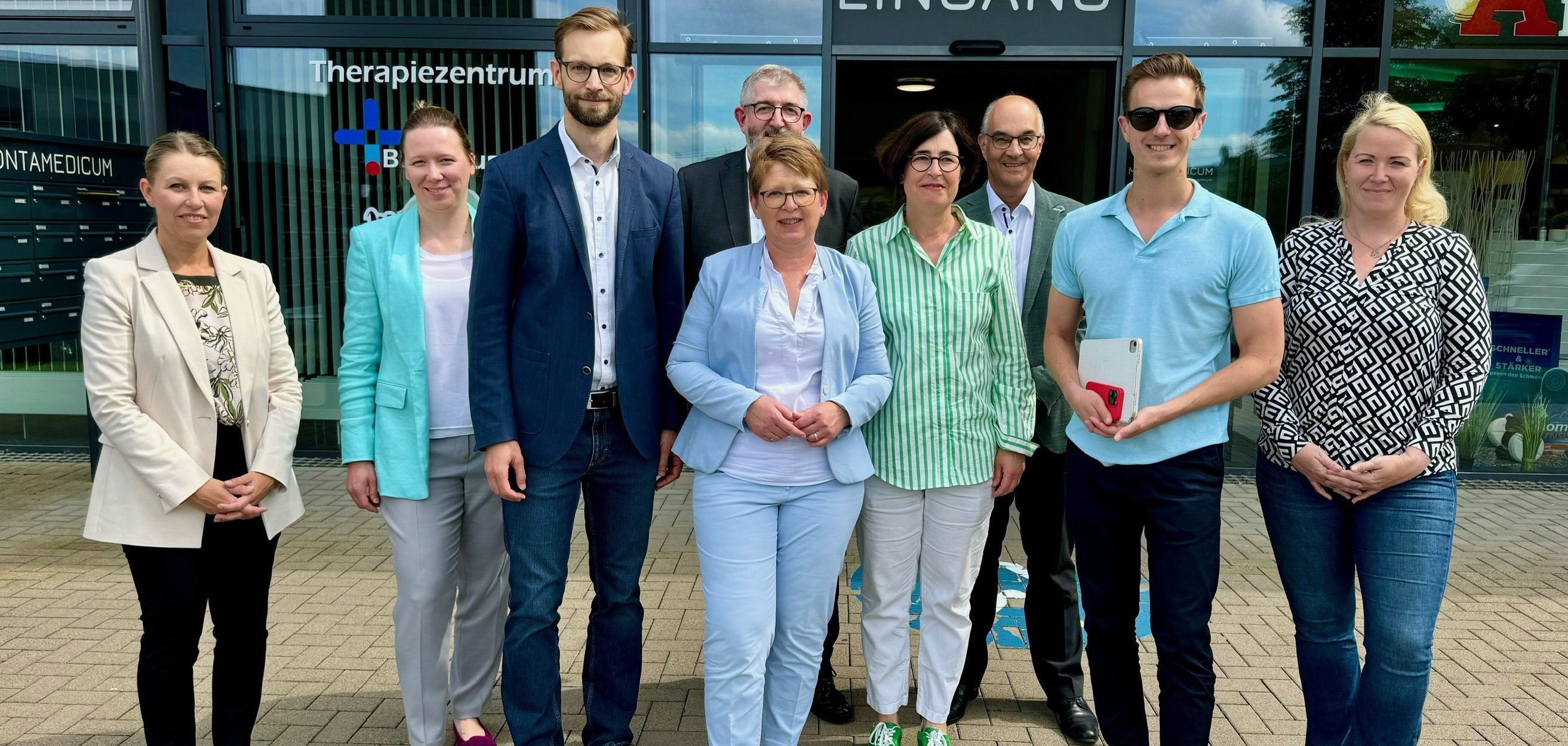 Eine Gruppe von neun Personen steht vor dem Eingang eines Therapiezentrums, darunter vier Frauen und fünf Männer. Die Gruppe trägt formelle bis halb-formelle Kleidung und posiert lächelnd für das Foto. Im Hintergrund sind die Schriftzüge "Therapiezentrum" und "Eingang" zu sehen.
