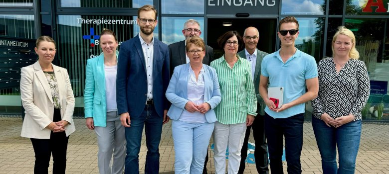 Eine Gruppe von neun Personen steht vor dem Eingang eines Therapiezentrums, darunter vier Frauen und fünf Männer. Die Gruppe trägt formelle bis halb-formelle Kleidung und posiert lächelnd für das Foto. Im Hintergrund sind die Schriftzüge "Therapiezentrum" und "Eingang" zu sehen.