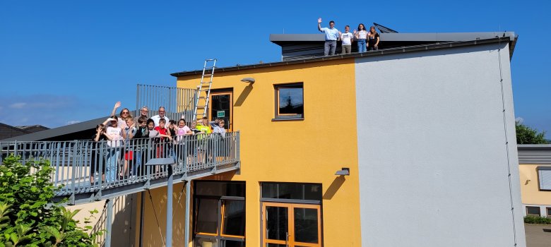 Eine Gruppe von Kindern und Erwachsenen steht auf einem Balkon und einer Dachterrasse eines gelb-grauen Gebäudes und winkt in die Kamera. Ein Leiter führt vom Balkon zum Dach, wo weitere Personen stehen und ebenfalls winken. Der Himmel ist klar und blau.