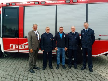 Fünf Männer stehen vor einem roten Feuerwehrfahrzeug mit der Aufschrift „Feuerwehr Nentershausen“. Drei der Männer tragen Feuerwehruniformen, einer trägt eine Jacke mit dem Feuerwehrwappen, und einer ist in einem Anzug gekleidet. Sie lächeln alle in die Kamera und posieren auf einem gepflasterten Platz.