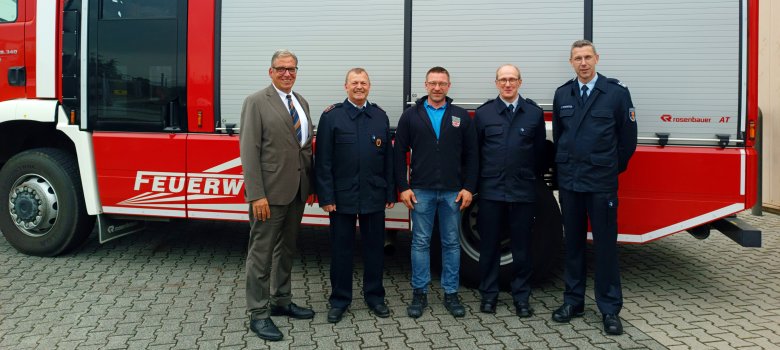 Fünf Männer stehen vor einem roten Feuerwehrfahrzeug mit der Aufschrift „Feuerwehr Nentershausen“. Drei der Männer tragen Feuerwehruniformen, einer trägt eine Jacke mit dem Feuerwehrwappen, und einer ist in einem Anzug gekleidet. Sie lächeln alle in die Kamera und posieren auf einem gepflasterten Platz.