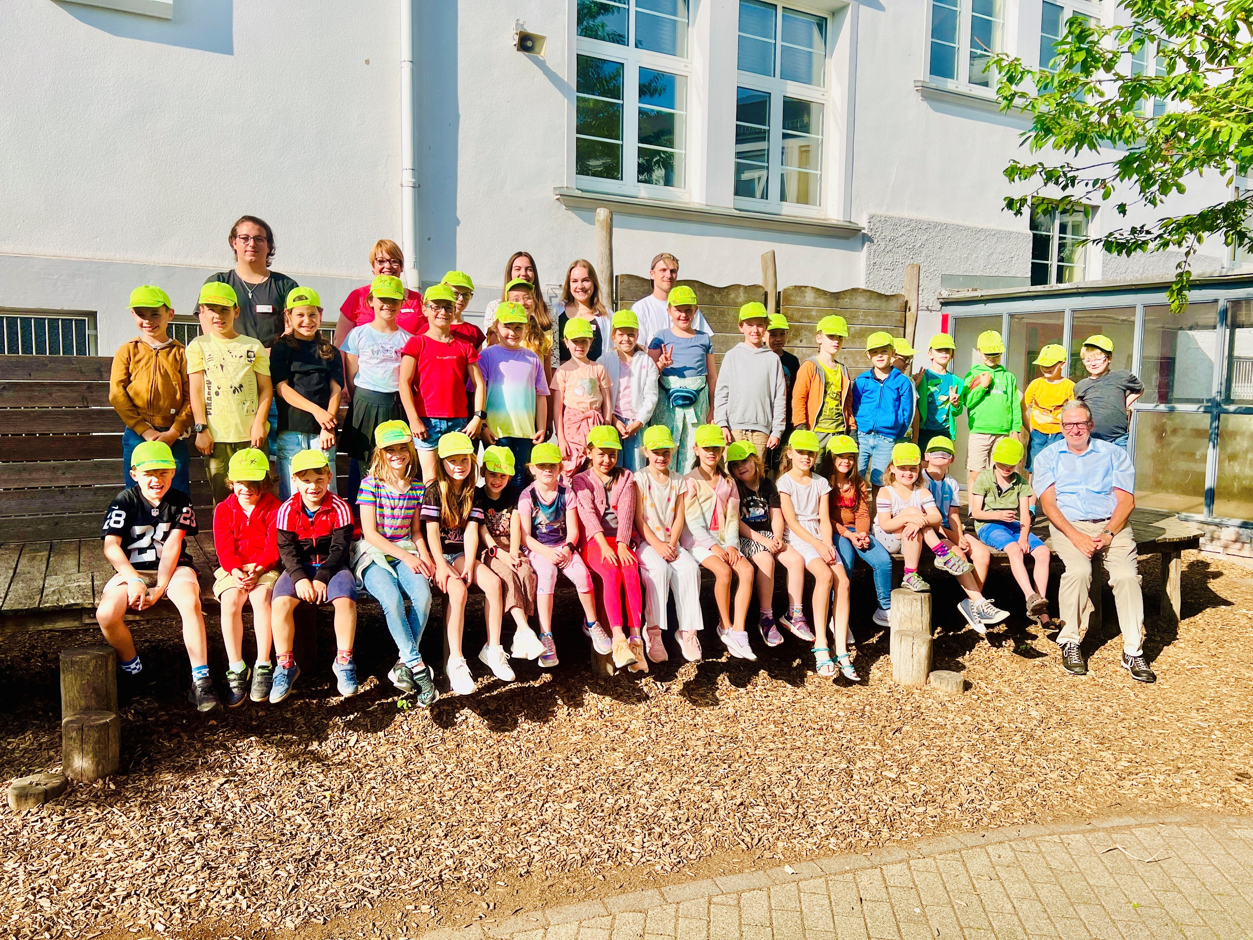 Eine große Gruppe von Kindern und Erwachsenen posiert für ein Foto im Freien vor einem weißen Gebäude mit Fenstern. Alle Kinder tragen gelbe Kappen und lächeln in die Kamera. Sie sitzen und stehen auf Holzbänken und Holzstufen, umgeben von Bäumen und hellem Sonnenschein.