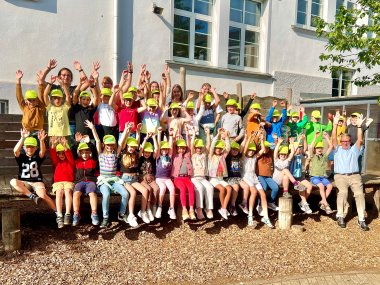 Eine große Gruppe von Kindern und Erwachsenen posiert vor einem weißen Gebäude für ein Foto im Freien. Alle Kinder tragen gelbe Kappen und heben jubelnd die Arme. Sie sitzen und stehen auf Holzbänken und Holzstufen, während die Sonne scheint und die Umgebung freundlich wirkt.