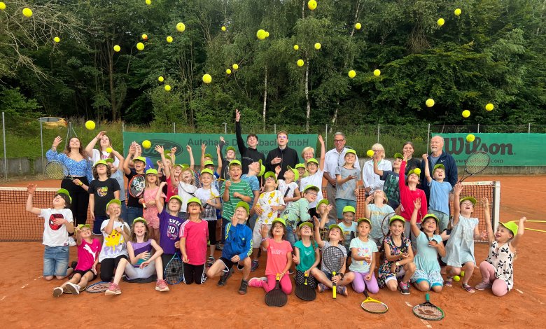 Eine große Gruppe von Kindern und Erwachsenen steht auf einem Tennisplatz und wirft gelbe Tennisbälle in die Luft. Alle Kinder tragen gelbe Kappen und viele halten Tennisschläger. Im Hintergrund sind grüne Bäume und ein Tennisnetz zu sehen. Die Stimmung ist fröhlich und lebhaft.