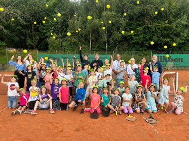 Eine große Gruppe von Kindern und Erwachsenen steht auf einem Tennisplatz und wirft gelbe Tennisbälle in die Luft. Alle Kinder tragen gelbe Kappen und viele halten Tennisschläger. Im Hintergrund sind grüne Bäume und ein Tennisnetz zu sehen. Die Stimmung ist fröhlich und lebhaft.