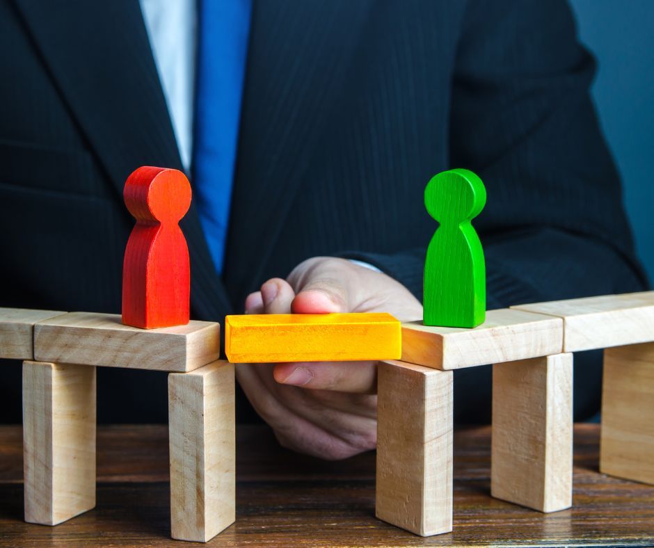 Eine Hand baut mit einem Bauklötzchen eine Brücke zwischen zwei Figuren.