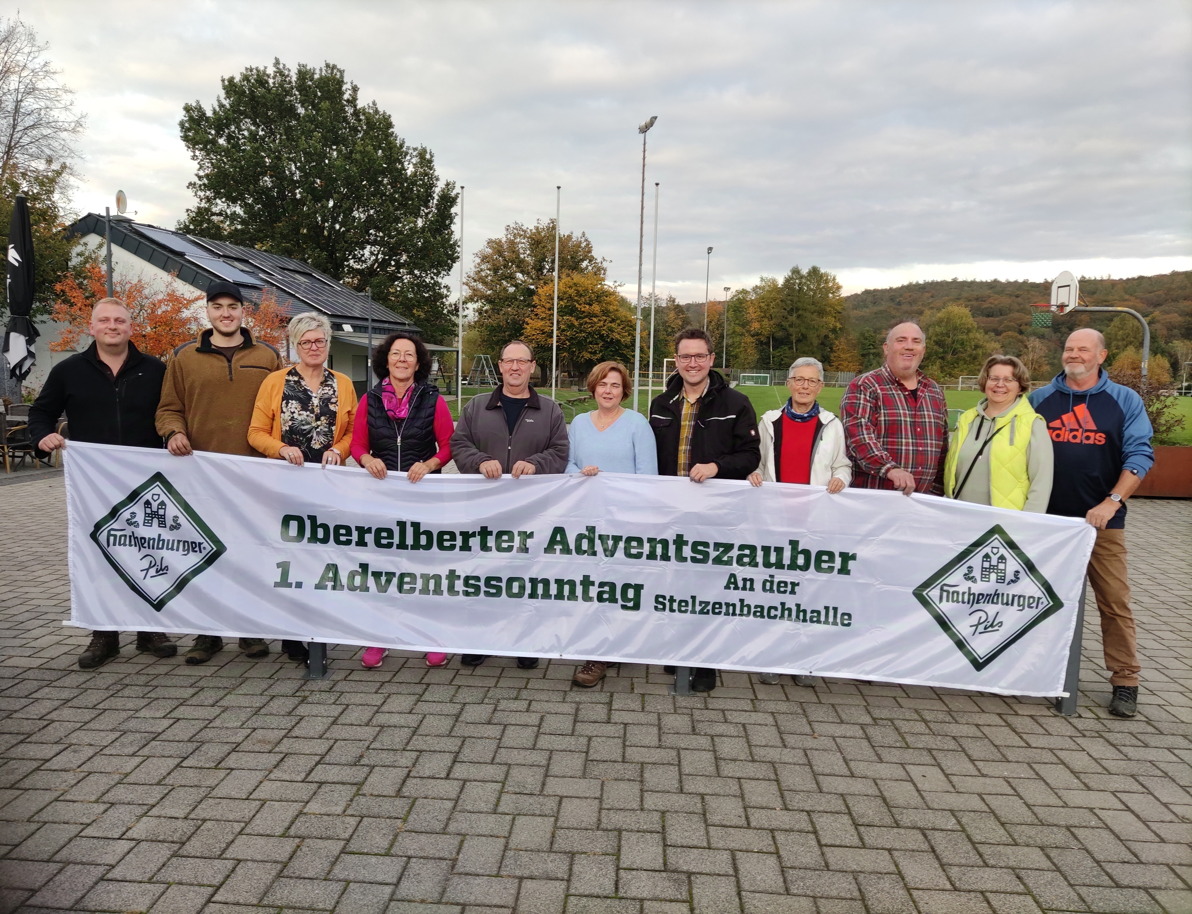 Eine Gruppe von elf Personen steht draußen und hält ein Banner mit der Aufschrift: „Oberelberter Adventszauber, 1. Adventssonntag an der Stelzenbachhalle“. Im Hintergrund sind ein Gebäude mit Solarpanels, herbstliche Bäume und ein Sportplatz zu sehen. Das Banner trägt die Logos von „Hachenburger Pils“.