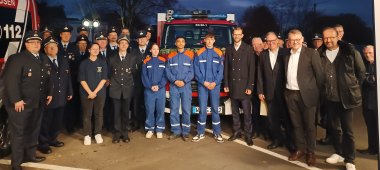 Eine Gruppe von Feuerwehrleuten und jungen Feuerwehranwärtern steht in Uniform vor einem Feuerwehrfahrzeug, begleitet von mehreren Personen in Zivilkleidung. Die Aufnahme scheint am Abend gemacht worden zu sein, vor einem dunklen Himmel, mit beleuchteten Feuerwehrfahrzeugen im Hintergrund. Alle blicken lächelnd in die Kamera, was auf eine feierliche Veranstaltung, möglicherweise die Übergabe eines neuen Fahrzeugs, hinweist.