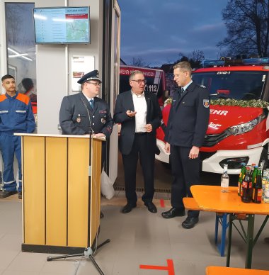 Drei Männer, zwei in Feuerwehruniform und einer in Zivilkleidung, stehen in einem Feuerwehrhaus nebeneinander und unterhalten sich neben einem Rednerpult. Im Hintergrund ist ein Feuerwehrfahrzeug zu sehen, während rechts im Bild Getränke auf einem Biertisch stehen. An der Wand hängt ein Bildschirm mit einer Karte.
