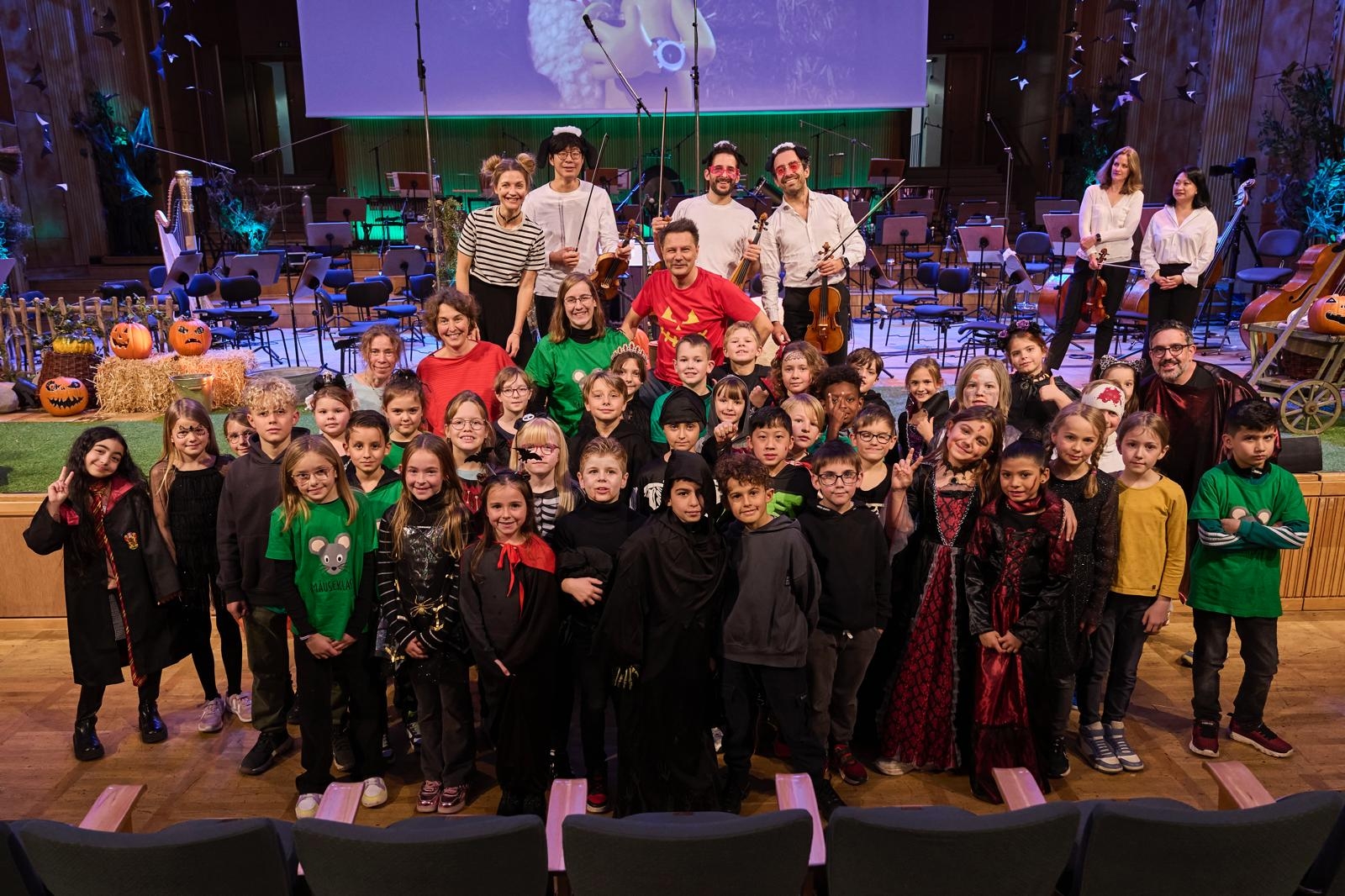 Eine Gruppe von Kindern in Halloween-Kostümen posiert mit Erwachsenen in einem Konzertsaal vor einer Bühne. Im Hintergrund sind ein Orchesteraufbau, Kürbisse, Strohballen und eine herbstlich dekorierte Kulisse zu sehen. Die Stimmung wirkt fröhlich und festlich, passend zum Anlass.