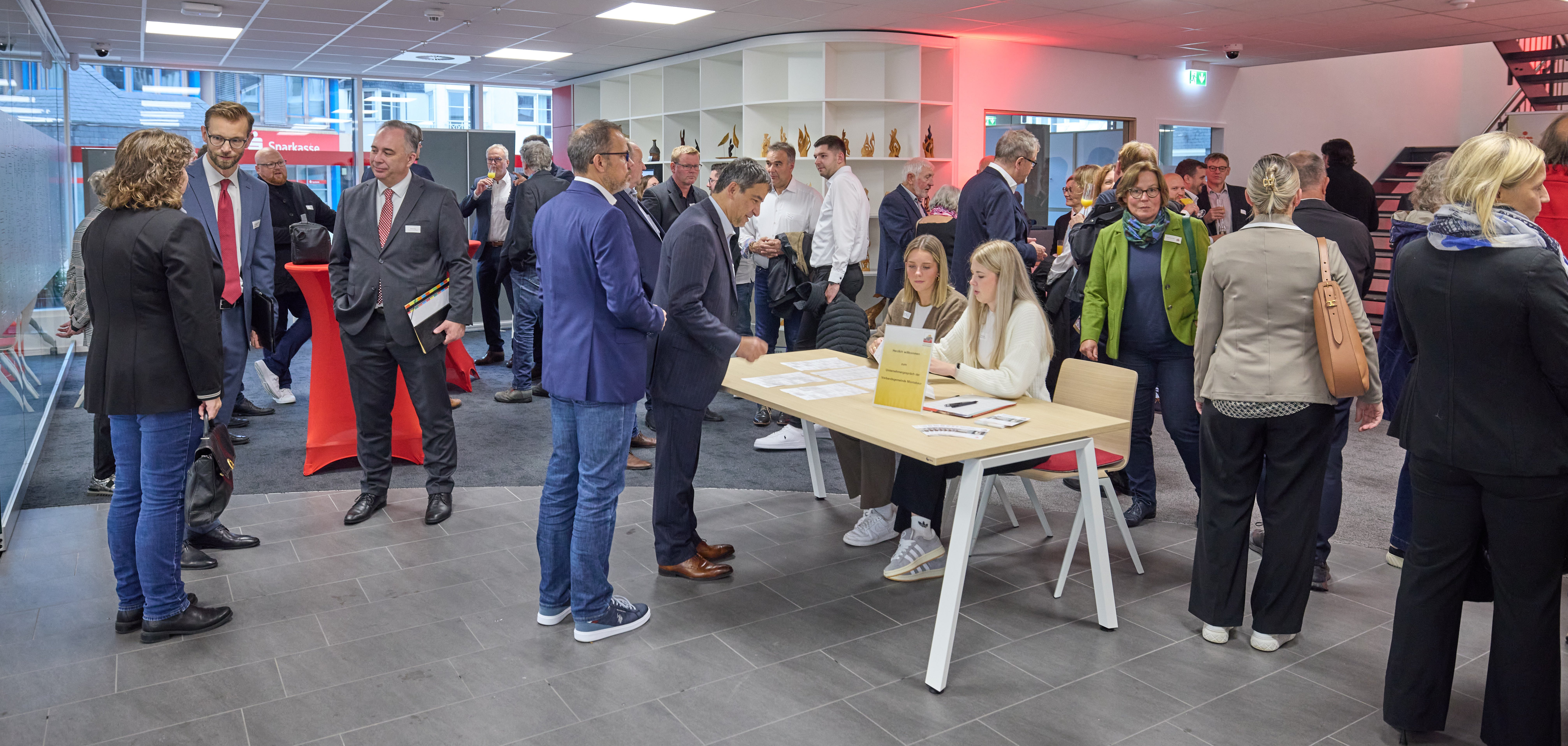 Menschen bei einer Netzwerkveranstaltung in einem modernen Raum mit Glaswand und heller Beleuchtung. Zwei Frauen sitzen an einem Tisch und begrüßen Gäste, während andere in Gruppen stehen und sich unterhalten. Im Hintergrund sind Regale mit Dekoration sowie ein roter Stehtisch zu sehen, was auf eine geschäftliche Atmosphäre hinweist.