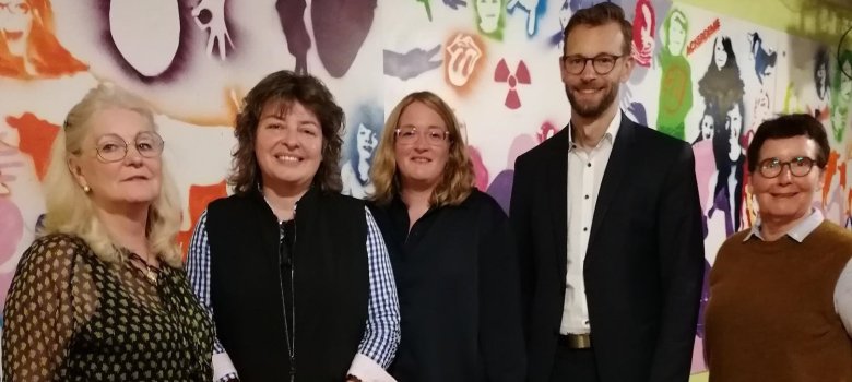 Fünf Personen stehen lächelnd in einer Reihe vor einer bunten Wand mit künstlerischen Symbolen und Silhouetten.