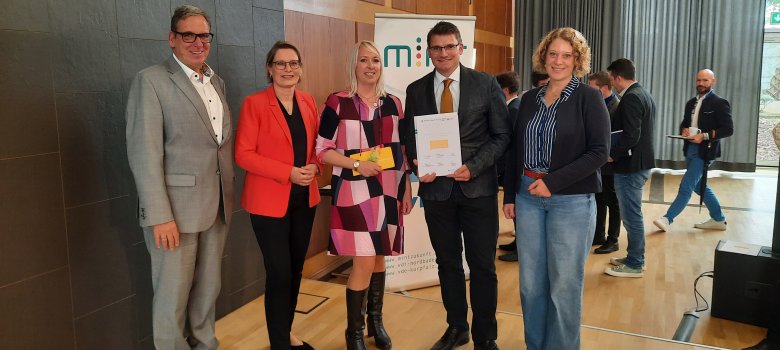 Eine Gruppe von fünf Personen steht lächelnd auf einer Bühne, offenbar während einer Preisverleihung. Zwei der Personen in der Mitte halten eine Auszeichnung oder Urkunde. Im Hintergrund unterhalten sich weitere Personen, während ein Mann von rechts ins Bild läuft. Die Atmosphäre wirkt förmlich und professionell.