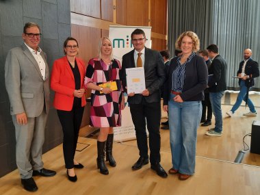 Eine Gruppe von fünf Personen steht lächelnd auf einer Bühne, offenbar während einer Preisverleihung. Zwei der Personen in der Mitte halten eine Auszeichnung oder Urkunde. Im Hintergrund unterhalten sich weitere Personen, während ein Mann von rechts ins Bild läuft. Die Atmosphäre wirkt förmlich und professionell.