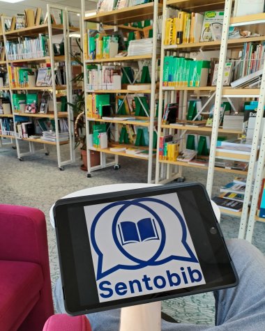 Ein Tablet wird in einem Raum mit Bücherregalen gehalten. Auf dem Bildschirm des Tablets ist das Logo von "Sentobib" zu sehen, das zwei Sprechblasen mit einem offenen Buch in der Mitte darstellt. Im Hintergrund befinden sich gut sortierte Bücher in einem hellen, gemütlichen Bereich einer Bibliothek.