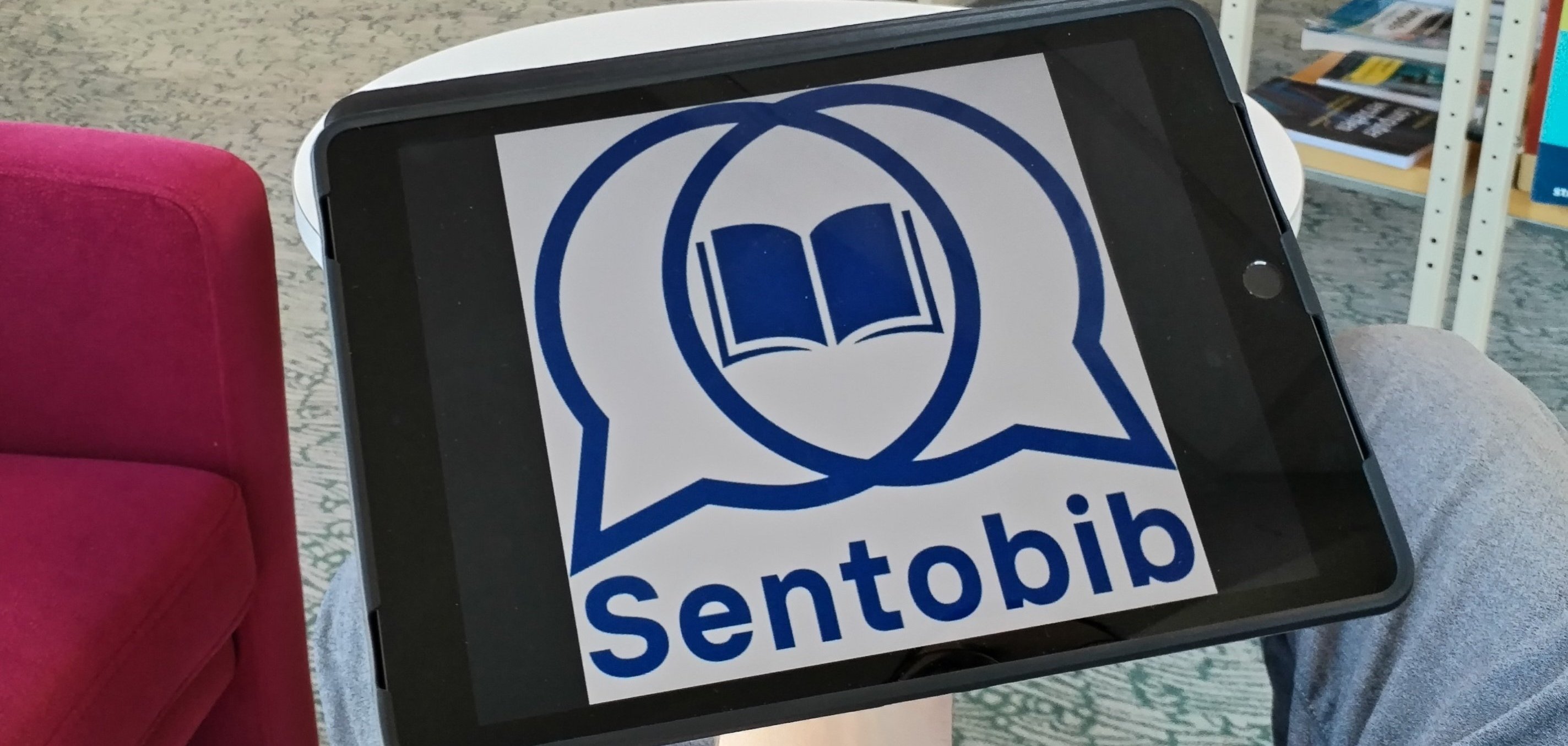 Ein Tablet wird in einem Raum mit Bücherregalen gehalten. Auf dem Bildschirm des Tablets ist das Logo von "Sentobib" zu sehen, das zwei Sprechblasen mit einem offenen Buch in der Mitte darstellt. Im Hintergrund befinden sich gut sortierte Bücher in einem hellen, gemütlichen Bereich einer Bibliothek.