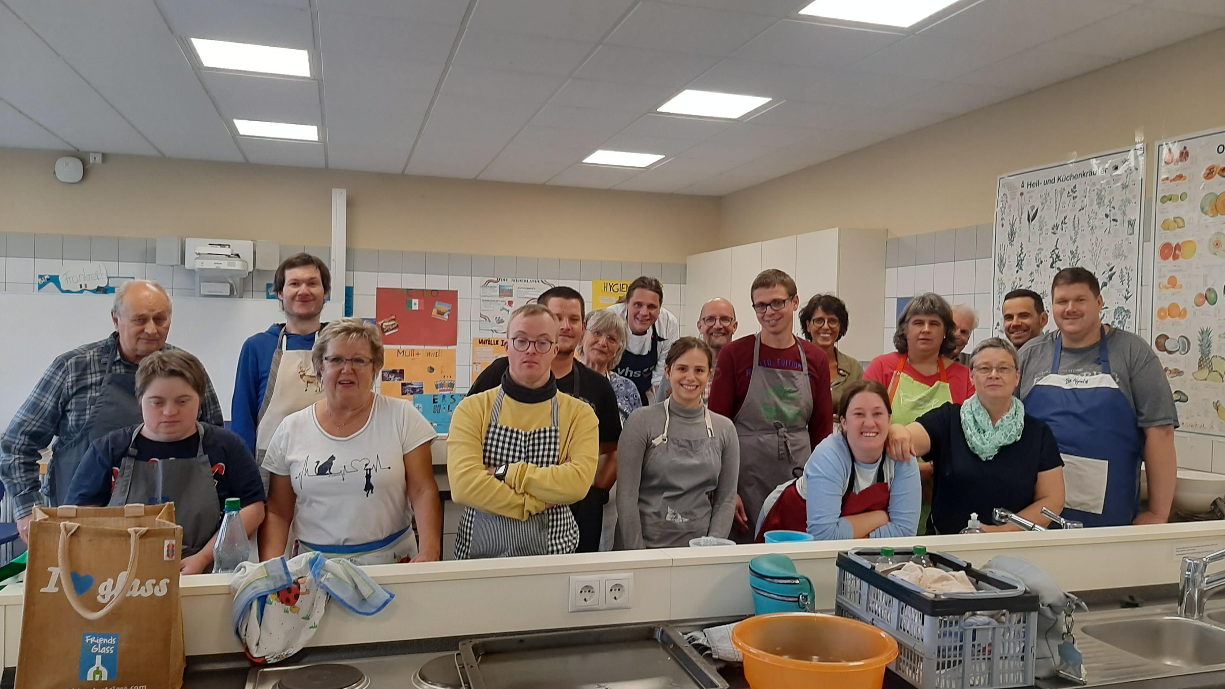 Das Bild zeigt eine Gruppe von Teilnehmenden eines inklusiven Tandem-Kochkurses, bei dem Menschen mit und ohne Behinderung gemeinsam kochen. Die fröhliche Atmosphäre im Kursraum deutet auf das gemeinsame Lernen und das Miteinander hin. Alle Teilnehmenden tragen Schürzen und stehen in einer Küche.