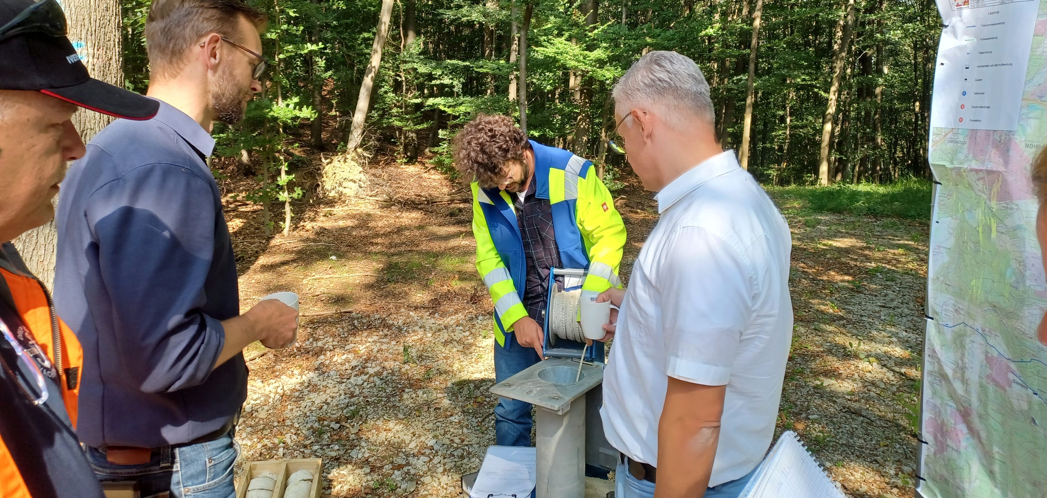 Mehrere Männer stehen im Wald um ein Bohrgerät herum, einer von ihnen führt eine Messung an dem Gerät durch. Einige der Männer halten Notizbücher und verfolgen den Prozess aufmerksam. Rechts im Bild hängt eine Landkarte an einem Gestell, und die Szene spielt sich unter sonnigen Bedingungen ab.