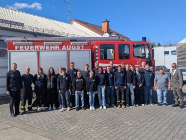 Gruppenfoto von etwa zwanzig Feuerwehrleuten und Ausbildern vor einem roten Einsatzfahrzeug mit der Aufschrift „FEUERWEHR AUGST“. Die Gruppe steht vor dem Feuerwehrhaus in Neuhausel bei sonnigem Wetter, im Hintergrund sind Gebäude und parkende Autos zu sehen. Alle Beteiligten wirken gut gelaunt und bereit für den Ausbildungsbeginn.