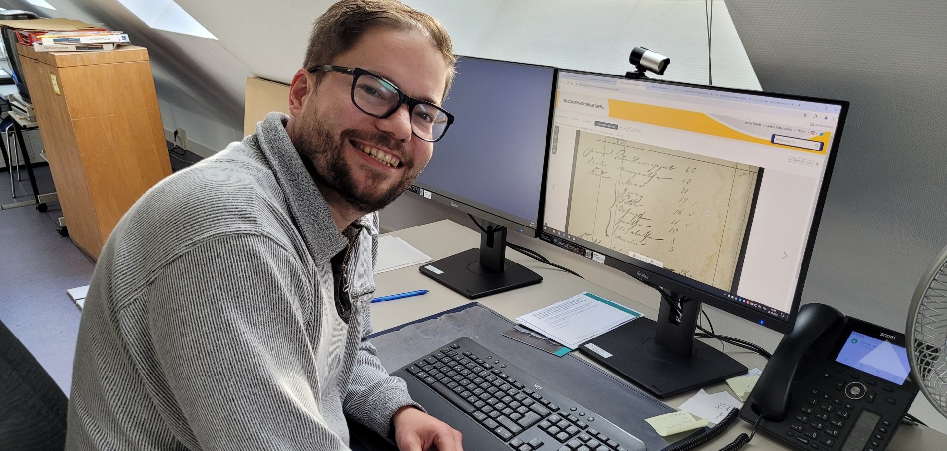 Ein lächelnder Mann mit Brille sitzt an einem Schreibtisch mit zwei Monitoren und arbeitet an der Digitalisierung historischer Dokumente. Auf dem Bildschirm ist eine handschriftliche Seite zu sehen, vermutlich aus einem Archiv. Der Arbeitsplatz ist modern eingerichtet mit Tastatur, Telefon und Notizzetteln.
