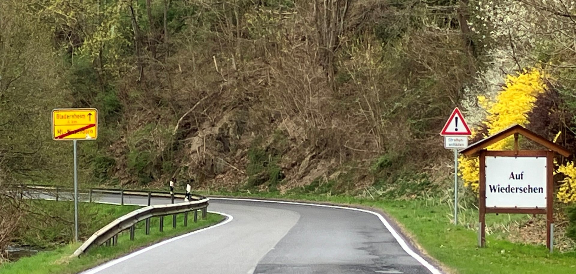 Ein kurviger Straßenabschnitt im ländlichen Gebiet mit mehreren Verkehrsschildern, darunter ein gelbes Ortsausgangsschild und ein großes Schild mit der Aufschrift "Auf Wiedersehen". Rechts neben der Straße blühen gelbe Forsythien, im Hintergrund ist ein bewaldeter Hang zu sehen.