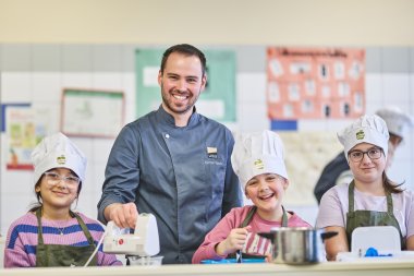Ein Koch im blauen Arbeitsoutfit steht lächelnd mit drei Kindern in Kochmützen und Schürzen in einer Schulküche. Die Kinder arbeiten mit Küchengeräten wie einem Handmixer und einem Kochtopf und wirken fröhlich und konzentriert beim gemeinsamen Kochen.