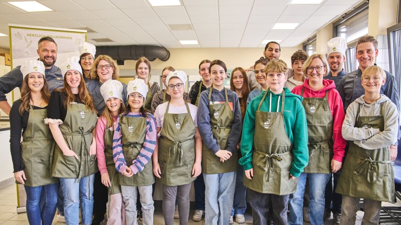 Eine große Gruppe aus Schülerinnen, Lehrerinnen und Köch*innen posiert fröhlich in einer Schulküche für ein Gruppenfoto. Alle tragen grüne Schürzen, einige auch weiße Kochmützen, und im Hintergrund ist ein Schild mit der Aufschrift „Herzlich Willkommen“ zu sehen.