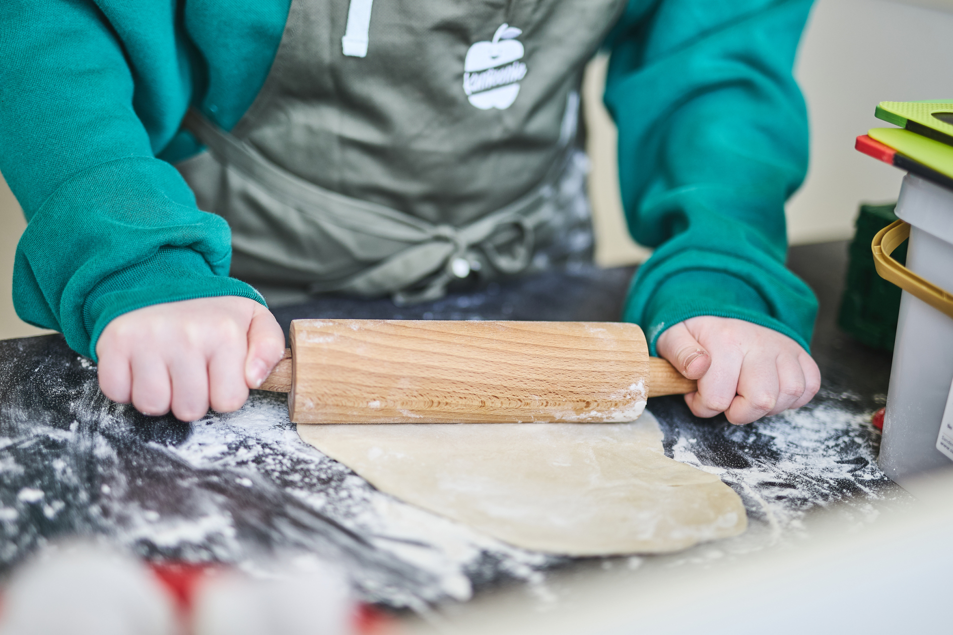 Ein Kind in grüner Kleidung und einer Küchenschürze rollt Teig mit einem hölzernen Nudelholz auf einer bemehlten Arbeitsfläche aus. Der Fokus liegt auf den Händen und dem Teig, während im Hintergrund Küchenutensilien zu sehen sind.