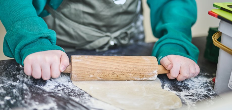 Ein Kind in grüner Kleidung und einer Küchenschürze rollt Teig mit einem hölzernen Nudelholz auf einer bemehlten Arbeitsfläche aus. Der Fokus liegt auf den Händen und dem Teig, während im Hintergrund Küchenutensilien zu sehen sind.