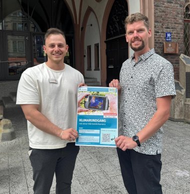 Zwei Männer stehen lächelnd vor einem historischen Gebäude mit gotischen Bögen und halten gemeinsam ein Plakat in die Kamera. Das Plakat bewirbt einen „Klimarundgang mit einer Wärmebildkamera“.
